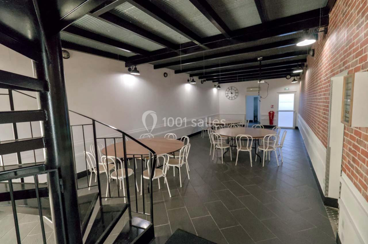 Salle avec tables rondes, chaises blanches, escalier en colimaçon au premier plan, murs en briques et éclairage suspendu.
