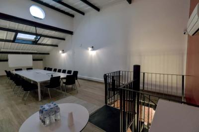 Salle avec des tables rondes, des chaises blanches, un mur en briques et un escalier en colimaçon métallique.