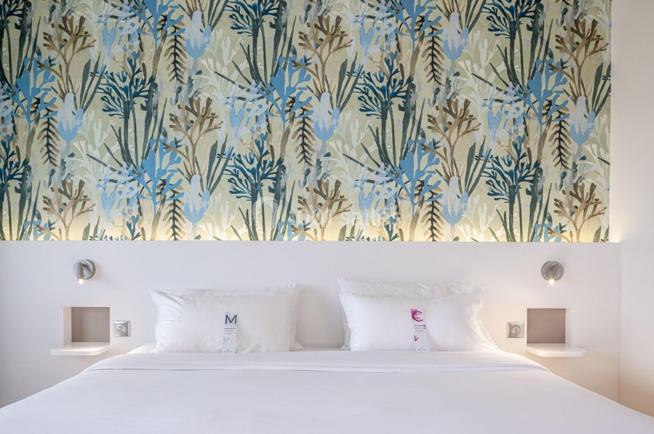 Lit double avec draps blancs, deux oreillers et une tête de lit éclairée, devant un mur décoré de motifs végétaux colorés.