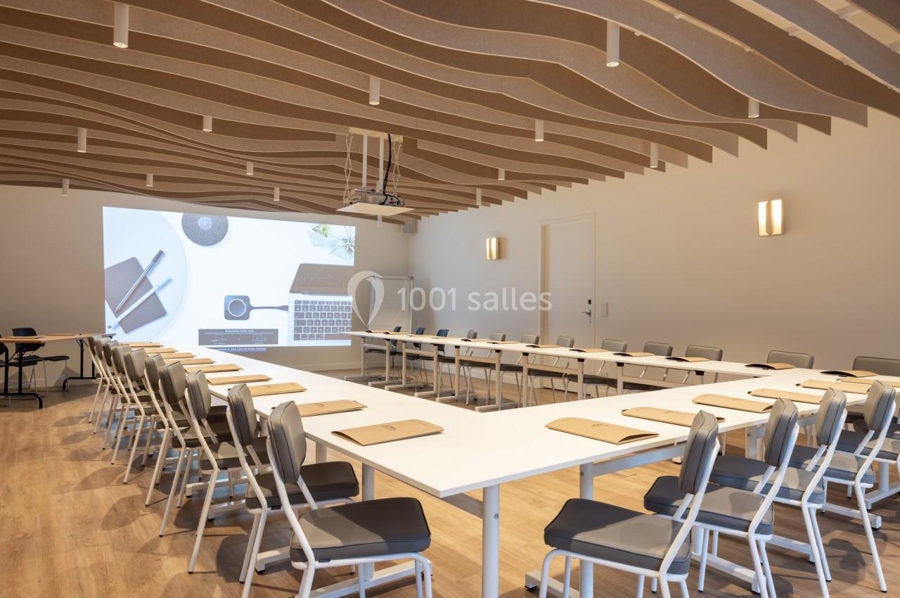 Salle de réunion moderne avec tables en U, chaises alignées, projecteur allumé et plafond au design ondulé.