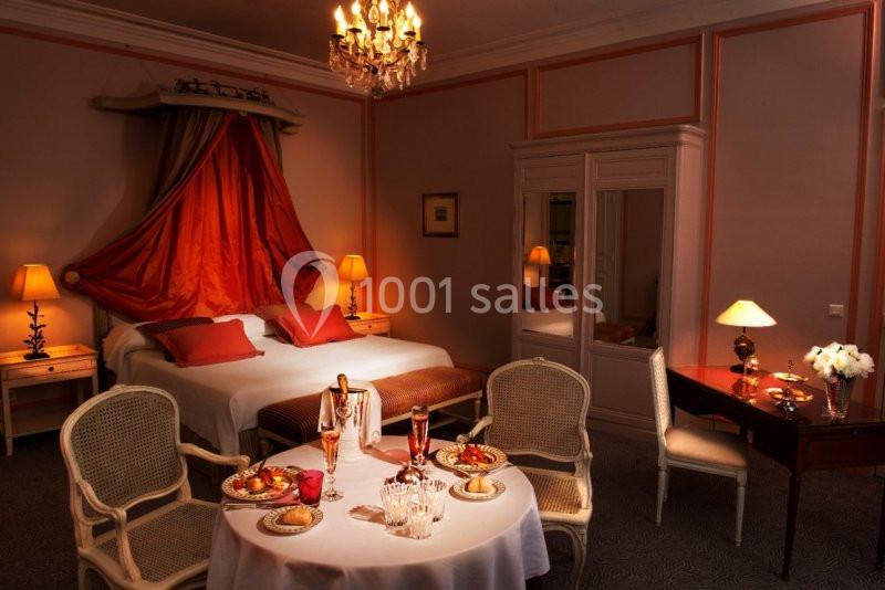 Chambre élégante avec lit double, baldaquin rouge, mobilier classique, table dressée et éclairage chaleureux.