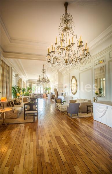 Salle lumineuse avec parquet en bois, canapés, miroirs dorés, grandes fenêtres et lustres imposants.