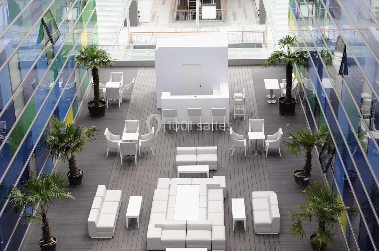 Terrasse moderne avec mobilier blanc, palmiers en pots et sols en bois, entourée de façades vitrées.