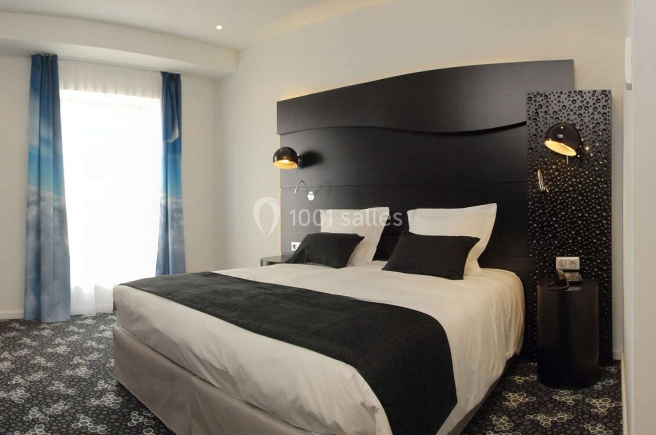 Chambre d'hôtel moderne avec lit double, tête de lit noire, lampes murales et rideaux bleus près d'une fenêtre.