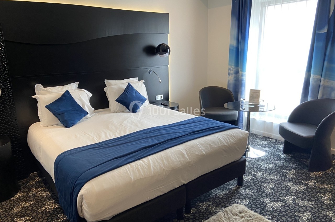 Chambre d'hôtel moderne avec lit double, draps blancs, coussins bleus, table ronde, deux fauteuils et grande fenêtre.