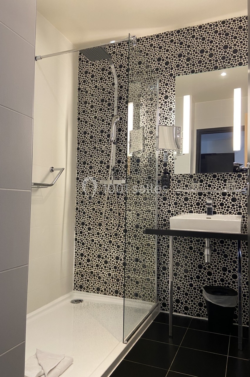 Salle de bain moderne avec douche à l'italienne, carrelage à motifs noirs et blancs, lavabo et miroir éclairé.