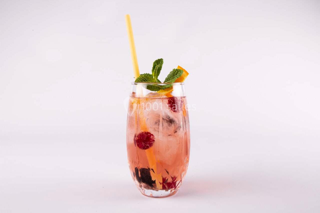 Verre de cocktail rose garni de glaçons, framboises, mûres, tranche d'orange, feuilles de menthe et une paille jaune.