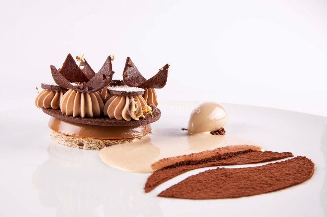 Dessert raffiné au chocolat avec mousse, éclats croustillants, crème et décorations dorées sur une assiette blanche.