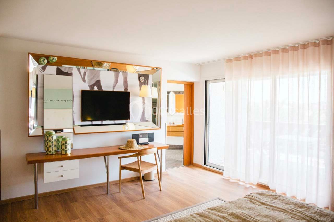 Chambre lumineuse avec un bureau en bois, un miroir mural, une télévision et des rideaux translucides près d'une baie vitrée.