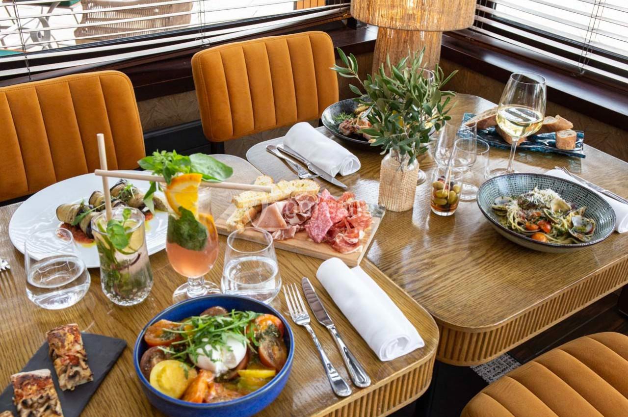 Table dressée avec des plats variés, boissons, couverts et décoration végétale dans un cadre chaleureux.