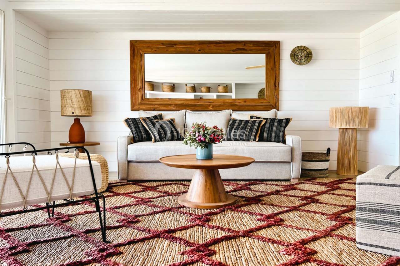 Salon lumineux avec canapé beige, coussins rayés, table basse en bois, tapis à motifs rouges et miroir encadré en bois.