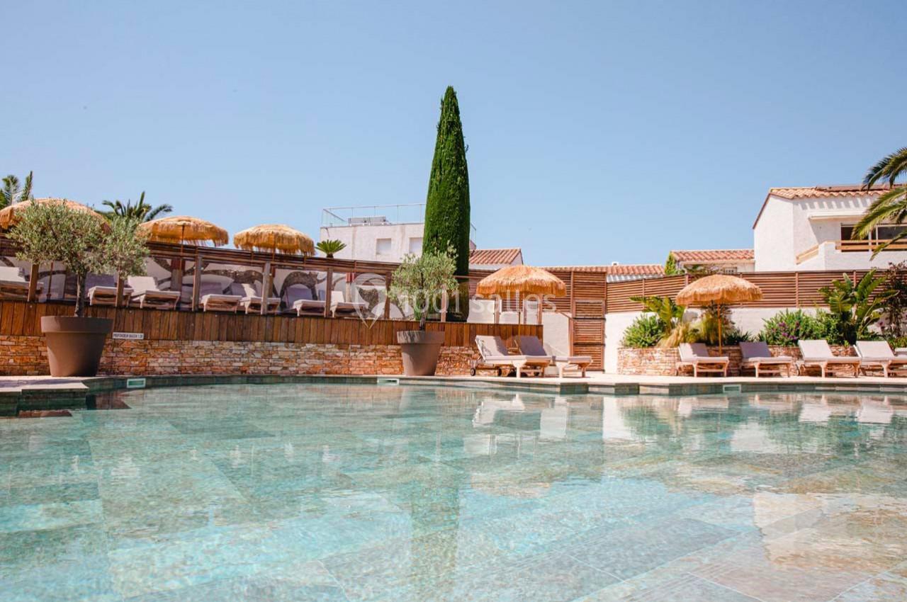 Piscine extérieure entourée de transats, parasols en paille, végétation et bâtiments aux murs clairs sous un ciel bleu.