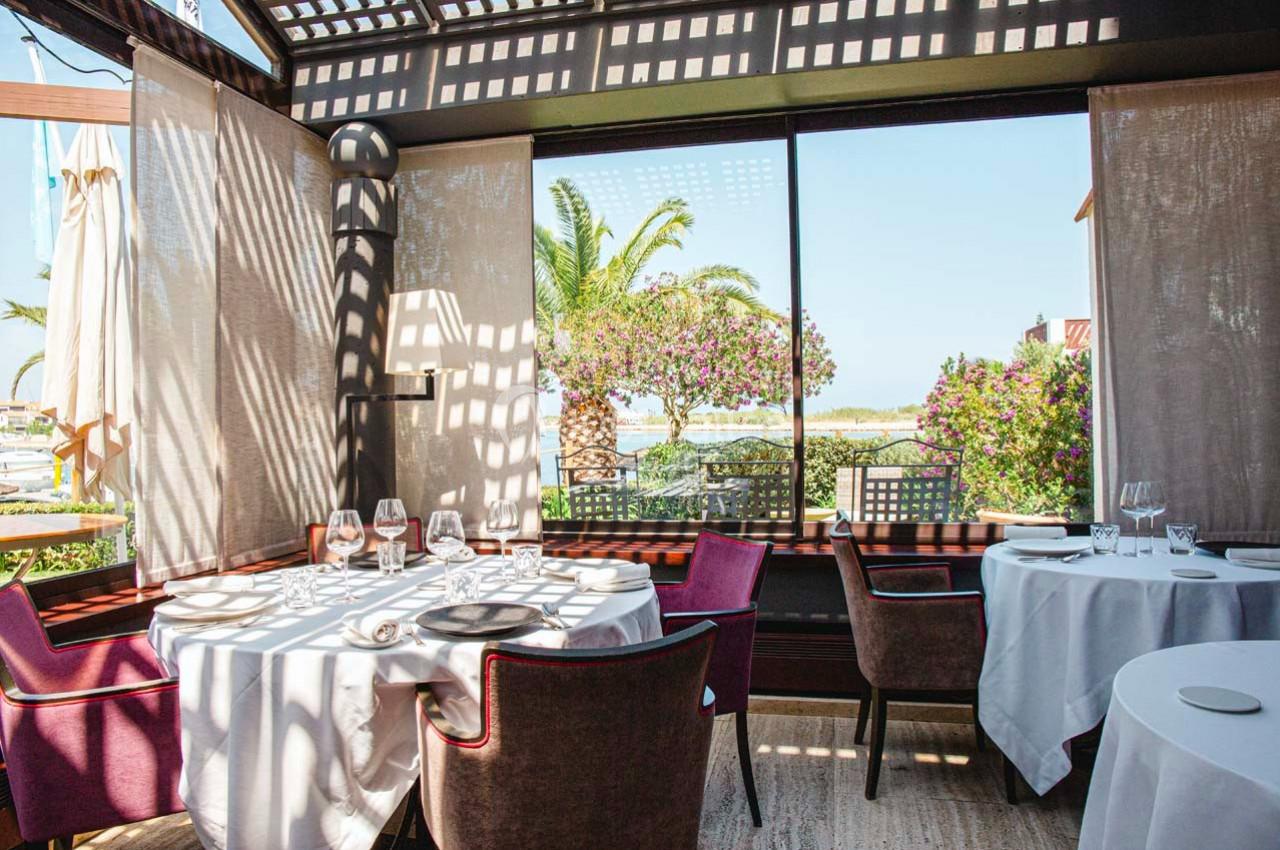 Salle de restaurant lumineuse avec vue sur un jardin fleuri, des palmiers et un paysage côtier en arrière-plan.