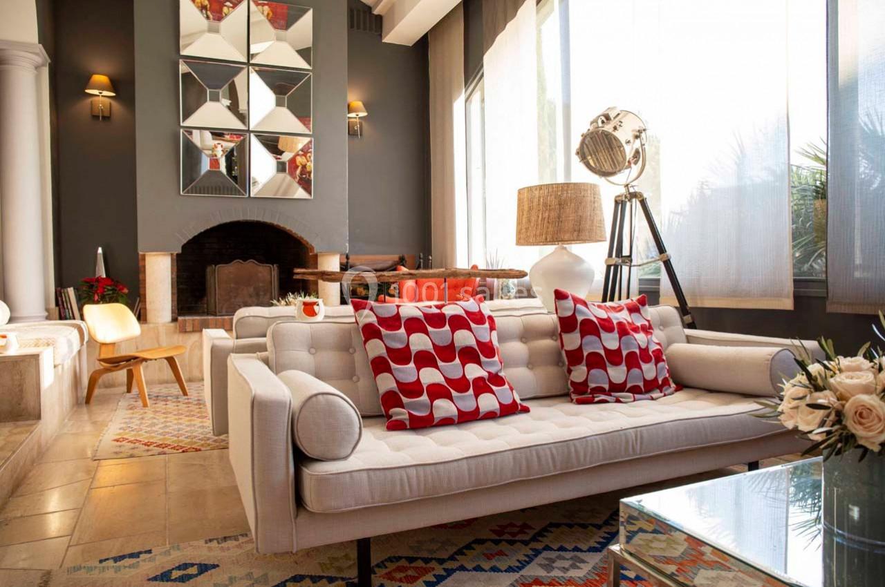 Salon moderne avec canapé blanc, coussins rouges à motifs, cheminée, lampadaire et grande fenêtre laissant entrer la lumière.