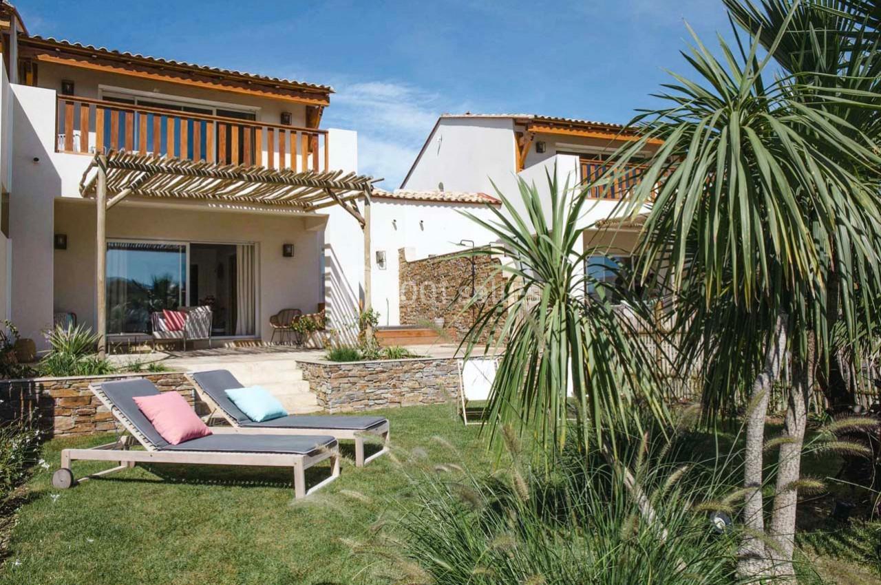 Jardin avec pelouse, transats, palmiers et terrasse ombragée devant une maison moderne à deux étages.
