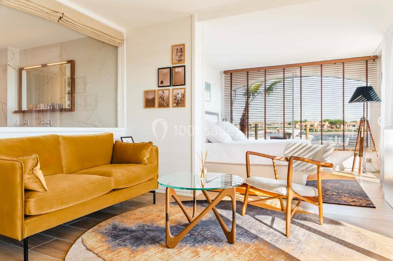 Salon lumineux avec canapé jaune, table basse en verre, fauteuils en bois et vue sur une chambre et l'extérieur.