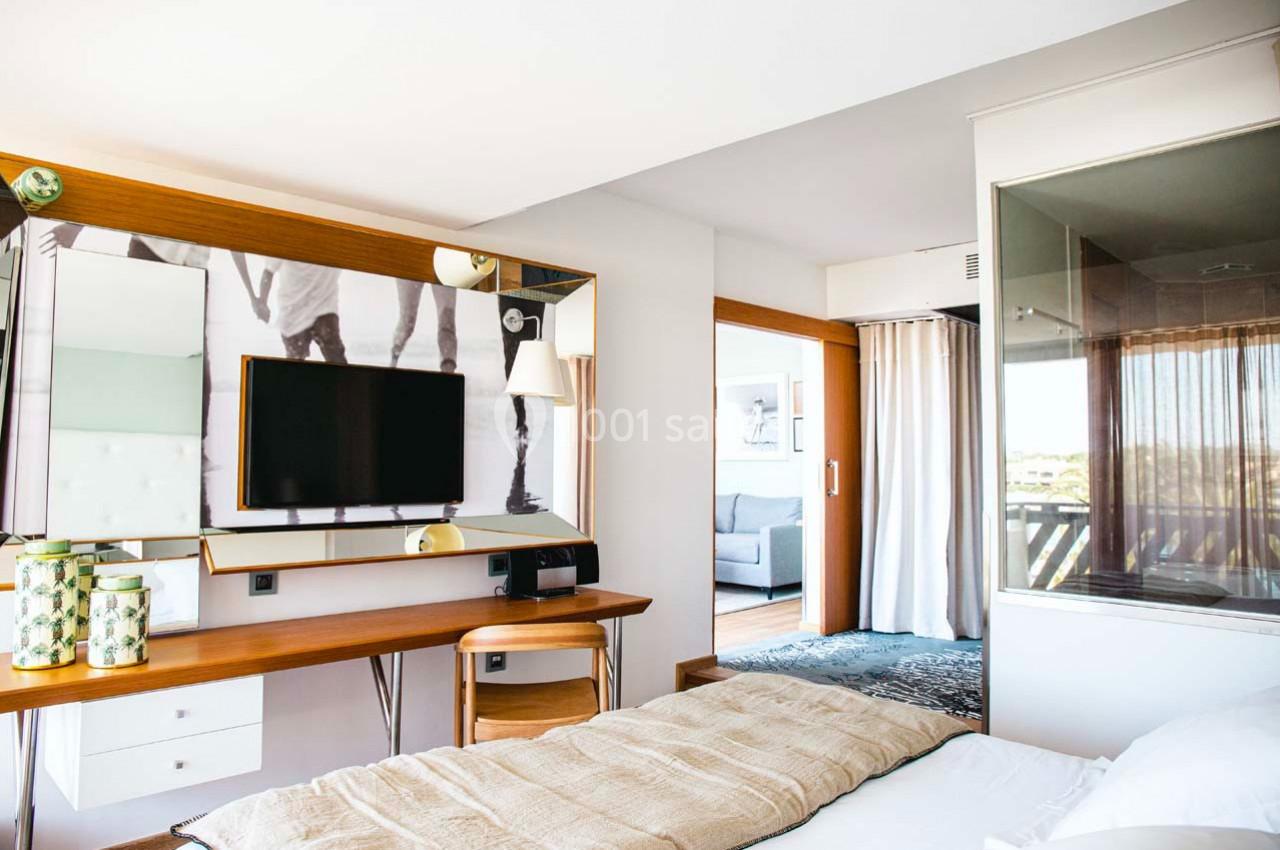 Chambre lumineuse avec lit, bureau en bois, télévision murale et vue sur un salon à travers une porte ouverte.