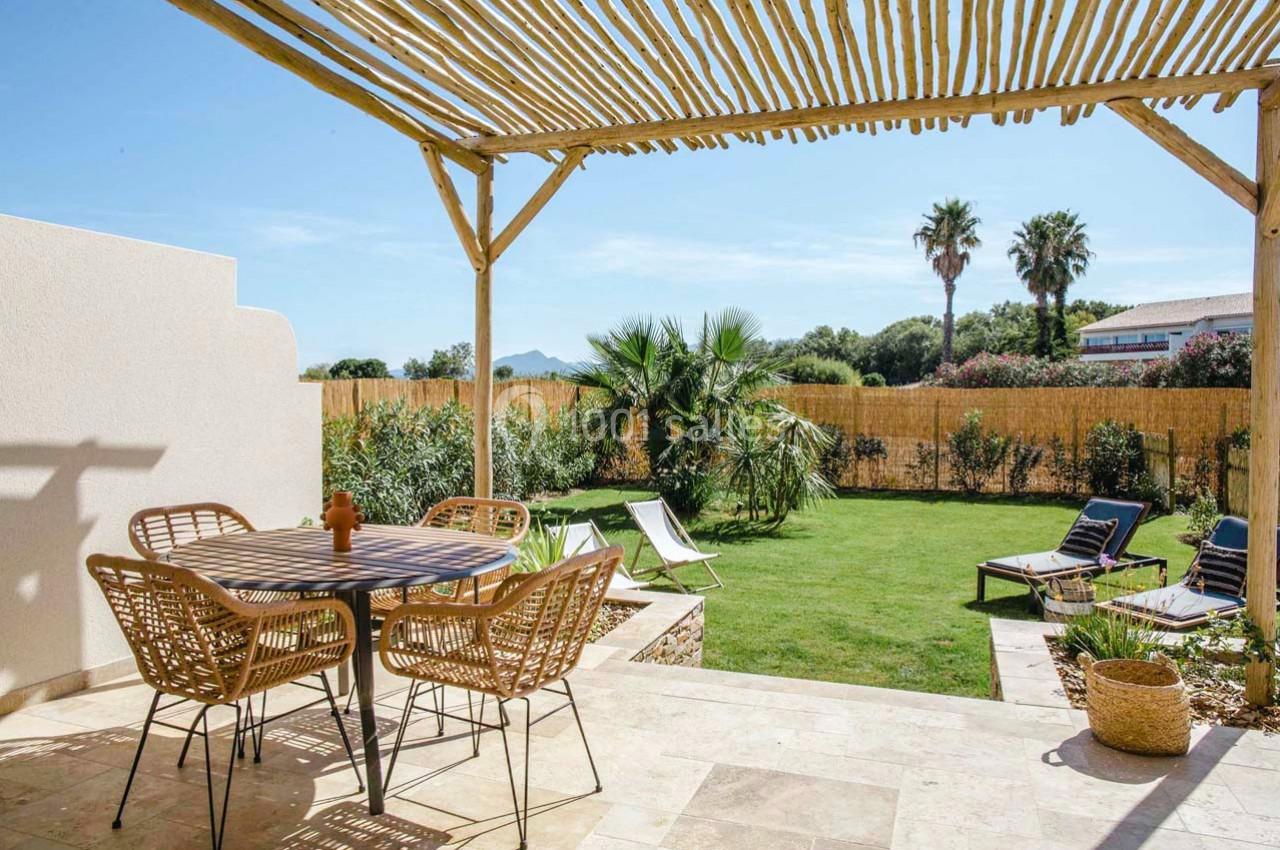 Terrasse avec table et chaises en rotin, pergola en bois, vue sur un jardin verdoyant avec palmiers et transats.
