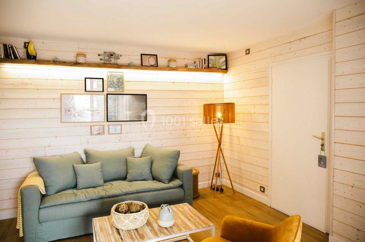 Salon lumineux avec murs en bois clair, canapé bleu, fauteuil jaune, lampe sur pied et étagère décorative.