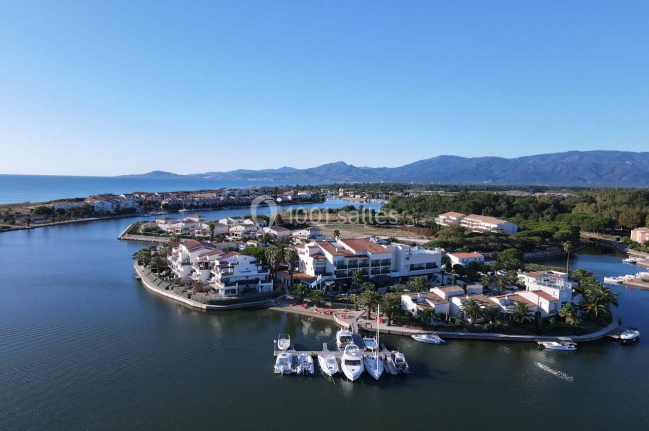 Vue aérienne d'une marina avec bateaux amarrés, entourée de bâtiments résidentiels et de montagnes en arrière-plan.