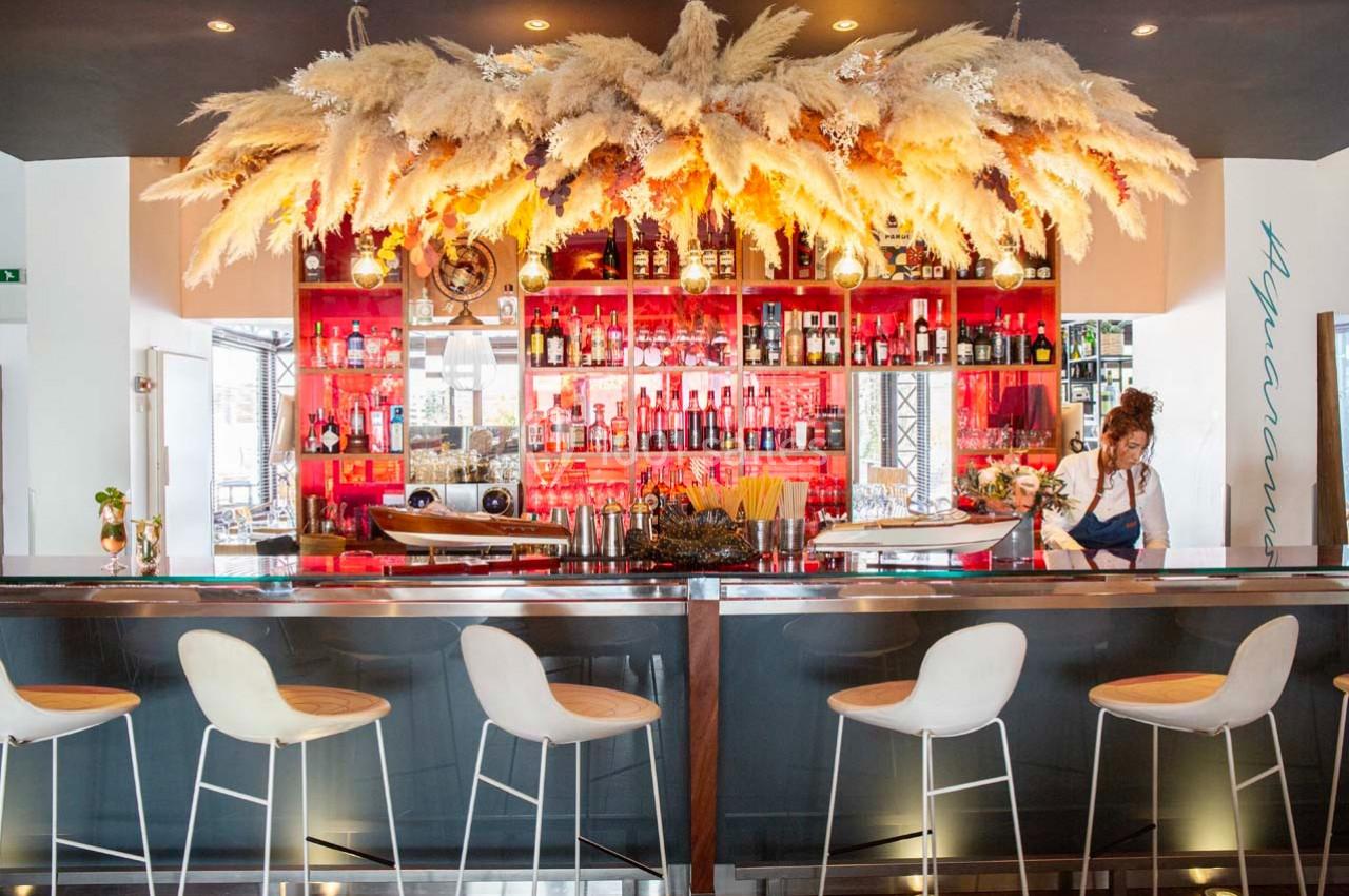 Bar moderne avec comptoir en métal, étagères rouges remplies de bouteilles et décoration suspendue en pampas.