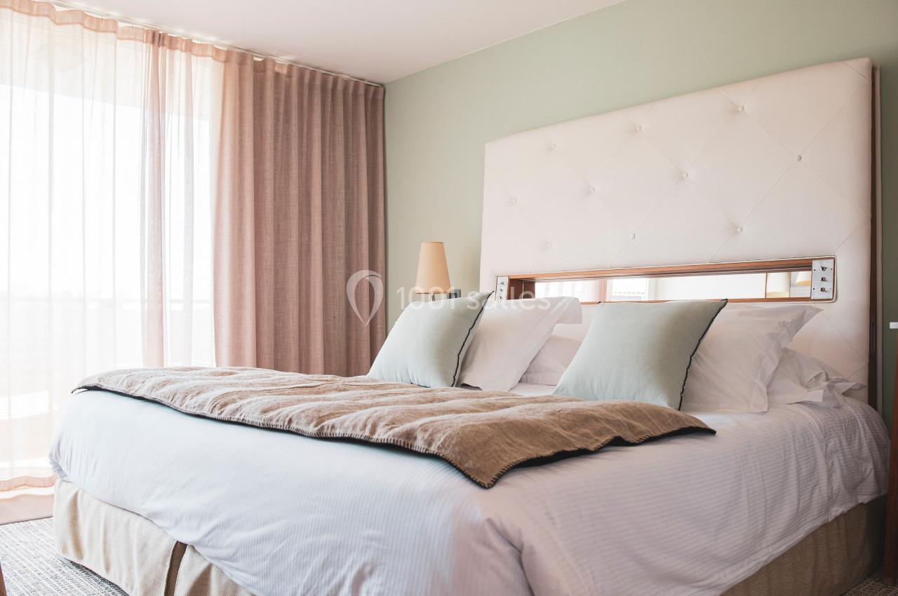 Lit double avec tête de lit capitonnée, draps blancs, coussins beiges, dans une chambre lumineuse aux rideaux roses.