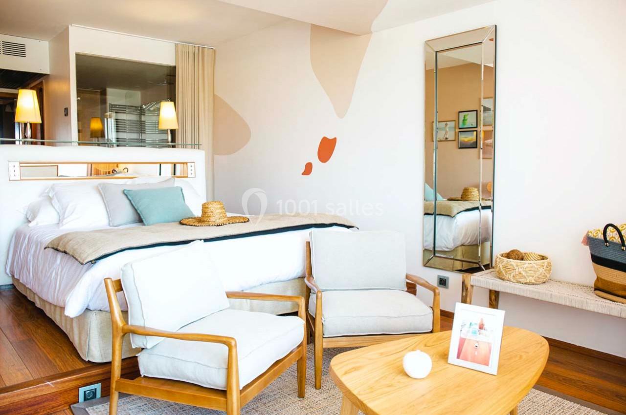 Chambre lumineuse avec lit double, fauteuils, table basse en bois, miroir mural et décoration moderne.