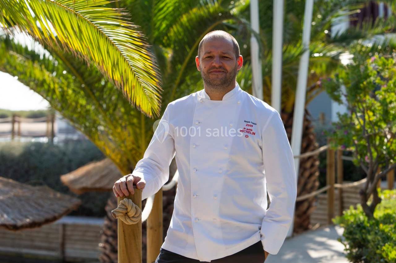 Un chef en veste blanche pose devant des palmiers et une terrasse ensoleillée.