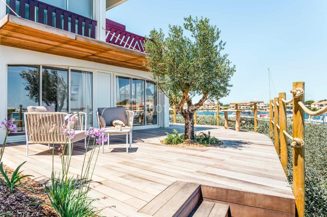 Terrasse en bois avec fauteuils, table basse, arbre et vue sur un port de plaisance en arrière-plan.