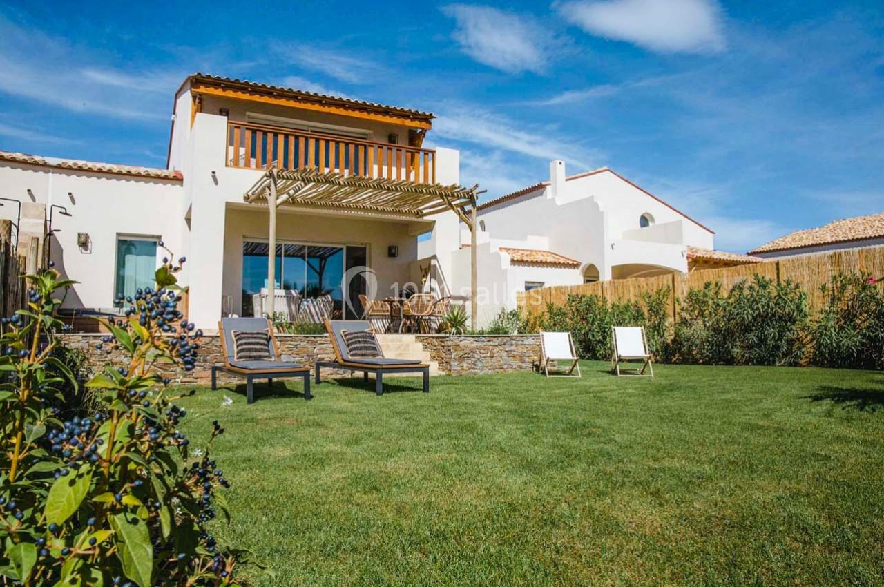 Maison moderne avec terrasse en bois, chaises longues et jardin verdoyant sous un ciel bleu.
