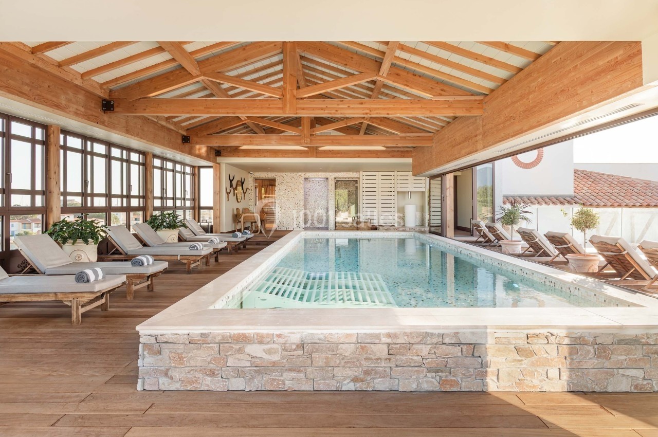 Piscine intérieure entourée de chaises longues, avec un plafond en bois et de grandes baies vitrées laissant entrer la…