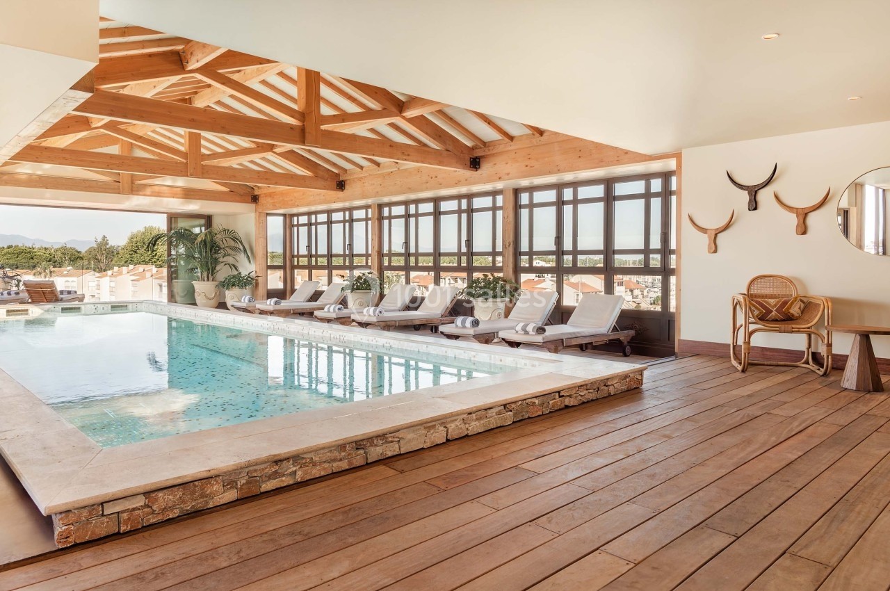 Piscine intérieure avec chaises longues, toit en bois, grandes fenêtres et vue sur des toits de maisons.