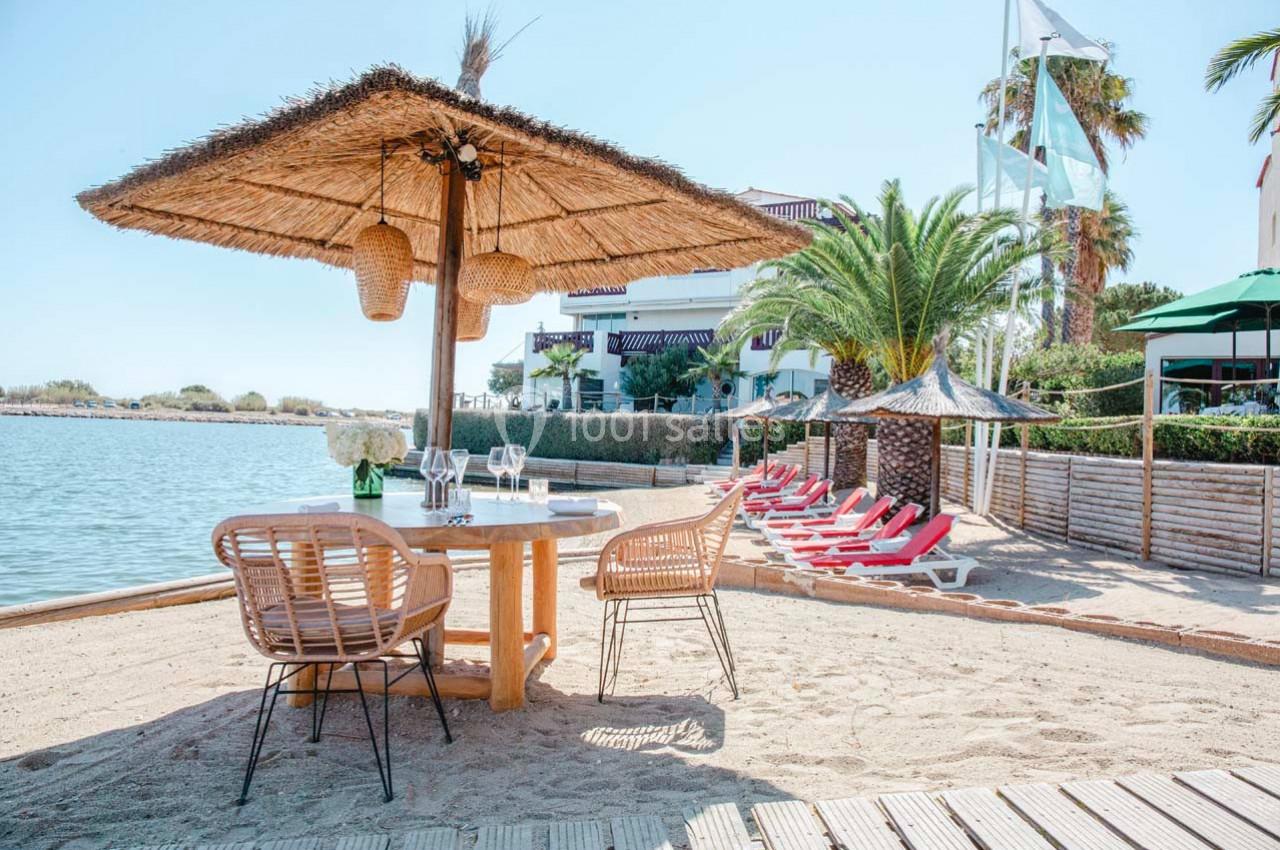 Table en bois avec chaises en rotin sous un parasol en paille, située sur une plage près d'un plan d'eau calme.