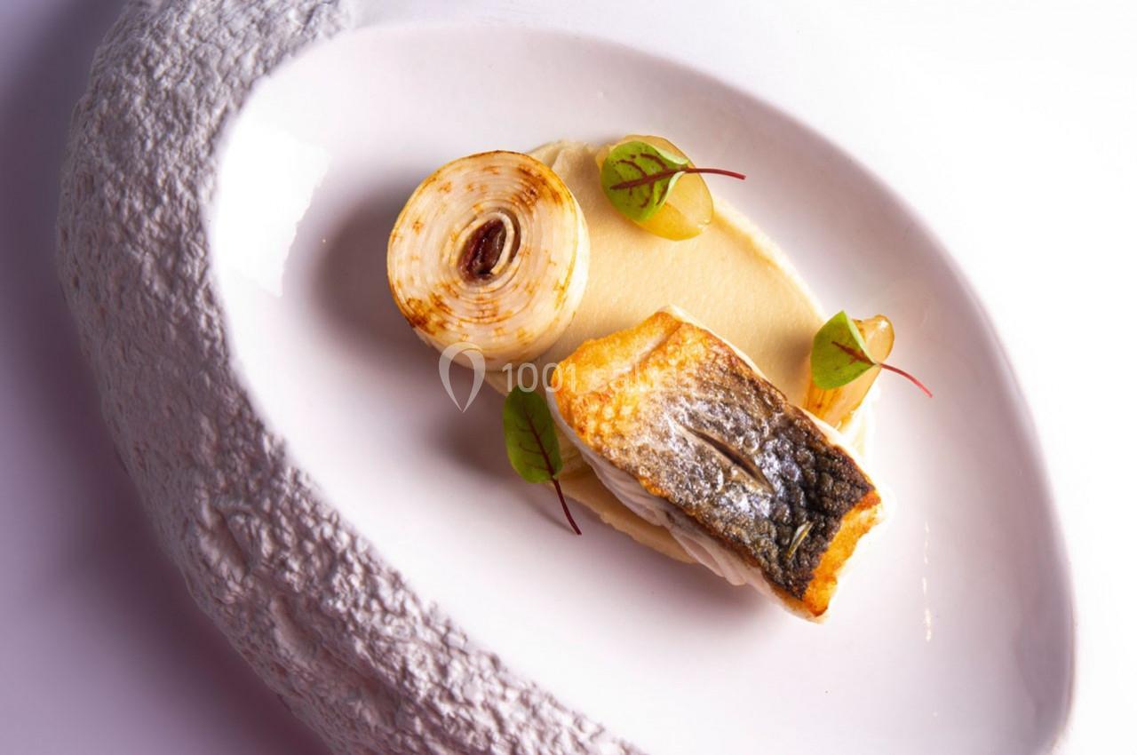 Filet de poisson grillé avec purée, oignon rôti et jeunes pousses, présenté sur une assiette blanche texturée.