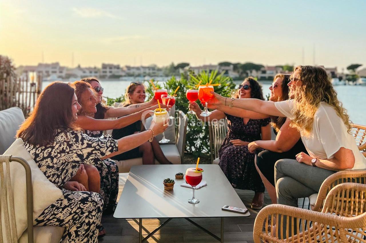 Un groupe de personnes trinque avec des cocktails en terrasse au bord de l'eau, sous une lumière de fin de journée.