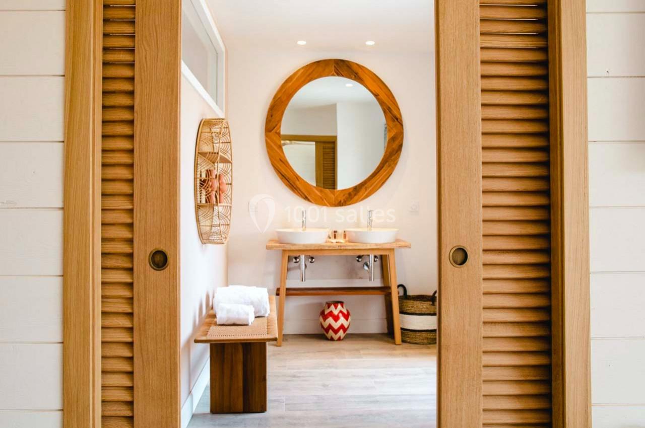 Salle de bain lumineuse avec deux vasques, miroir rond en bois, banc avec serviettes et éléments décoratifs.