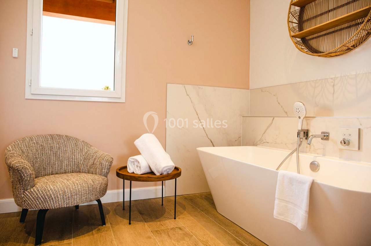 Salle de bain avec baignoire blanche, chaise en rotin, table en bois avec serviettes pliées et mur beige.