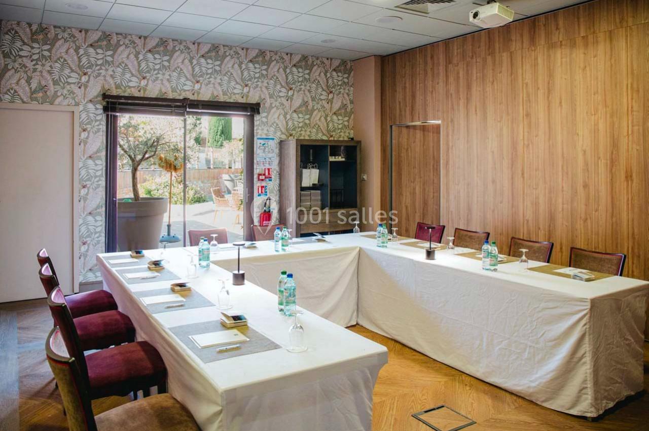Salle de réunion lumineuse avec tables disposées en U, chaises rouges, murs en bois et vue sur un jardin extérieur.