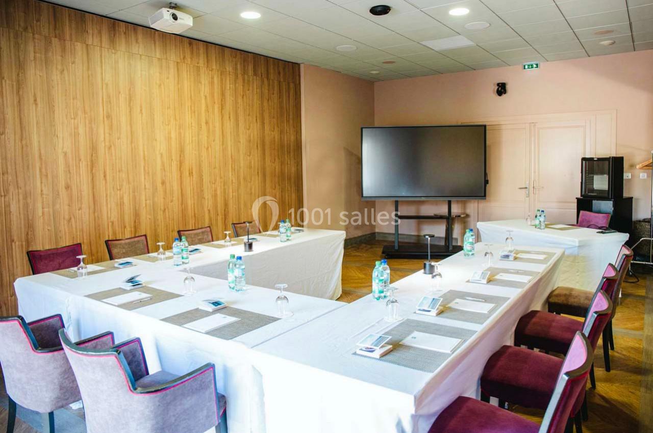 Salle de réunion lumineuse avec tables disposées en U, écran de présentation, chaises et bouteilles d'eau.
