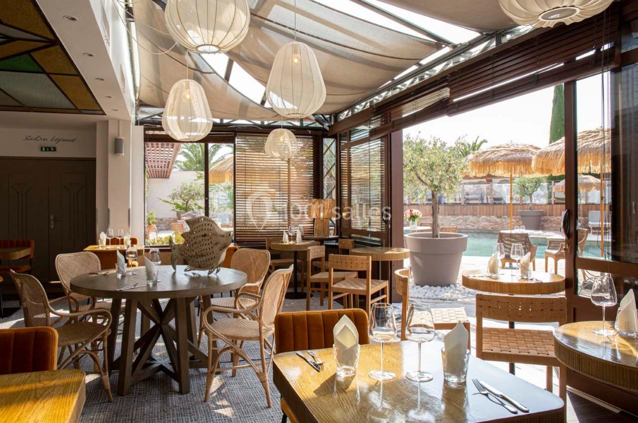Salle de restaurant lumineuse avec tables en bois, chaises tressées et vue sur une terrasse ensoleillée.