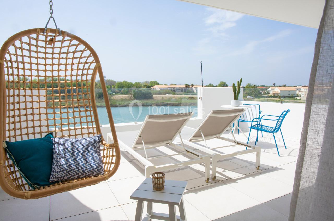 Terrasse avec vue sur un plan d'eau, équipée de transats, une chaise suspendue et une table avec des chaises.