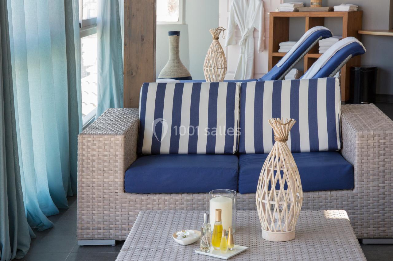 Salon lumineux avec canapé en rotin, coussins rayés bleus, table basse décorée et peignoirs accrochés en arrière-plan.