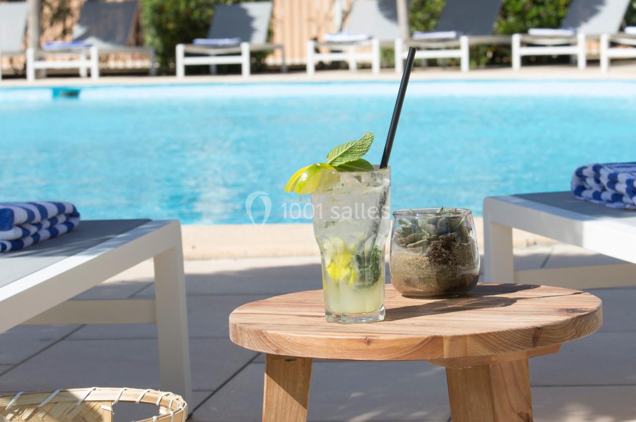 Verre de cocktail et plante sur une table en bois près d'une piscine entourée de transats et parasols.