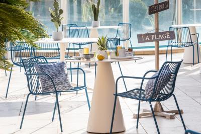 Terrasse ensoleillée avec tables et chaises bleues, décorée de plantes et d'un panneau indiquant ’La plage’.
