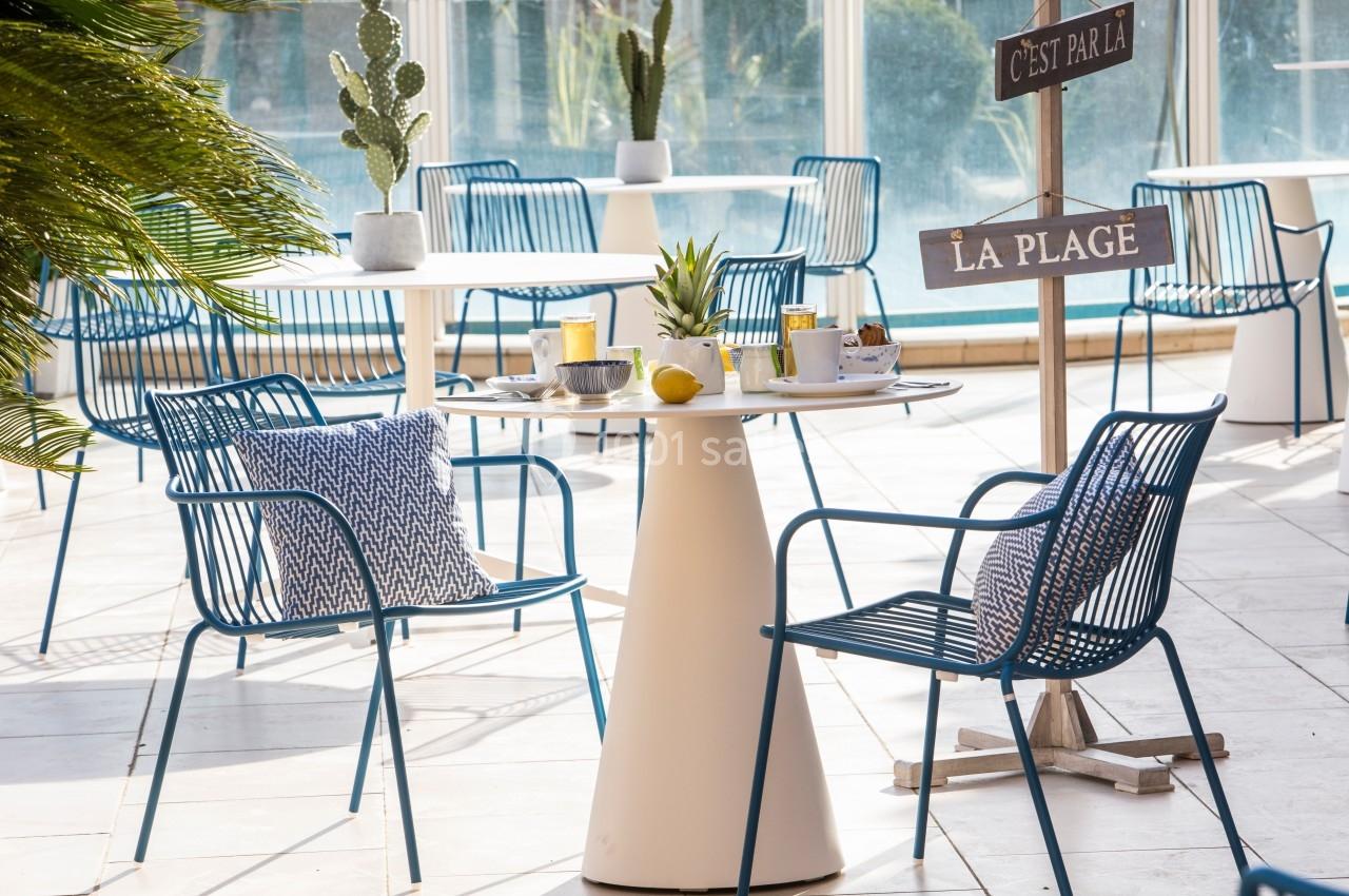 Terrasse ensoleillée avec tables et chaises bleues, décorée de plantes et d'un panneau indiquant ’La plage’.