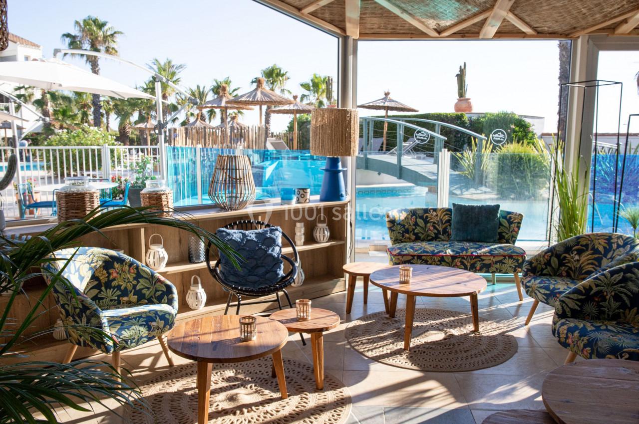 Espace lounge lumineux avec mobilier en bois et vue sur une piscine entourée de palmiers et parasols en paille.