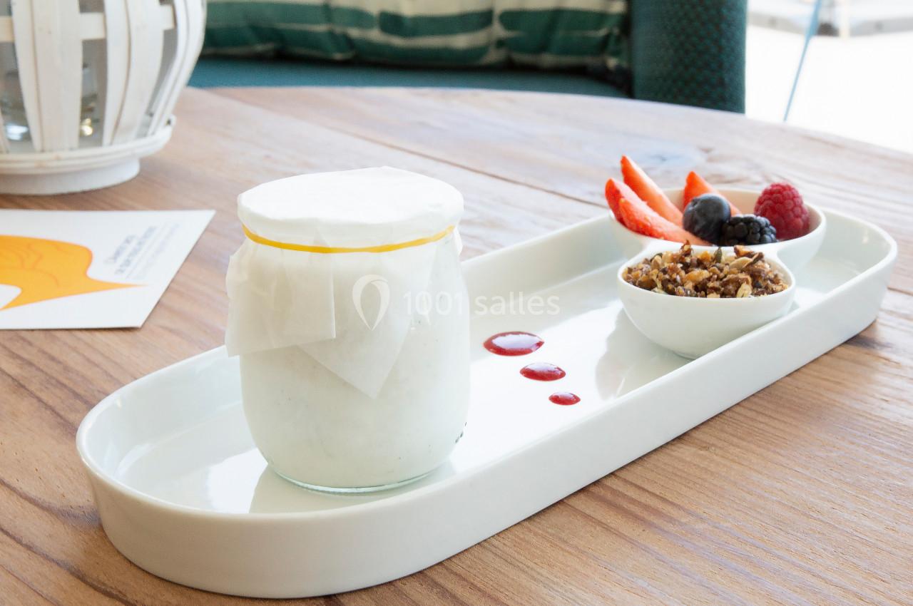 Pot de yaourt couvert de papier, accompagné de fruits rouges, granola et coulis sur un plateau blanc.