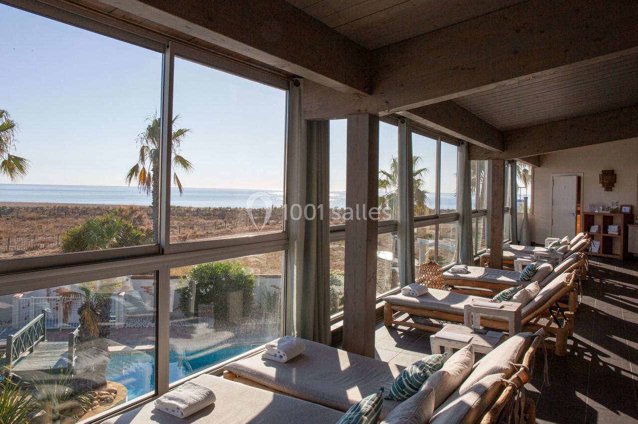 Salle lumineuse avec grandes baies vitrées, chaises longues alignées, vue sur une piscine, des palmiers et la mer.