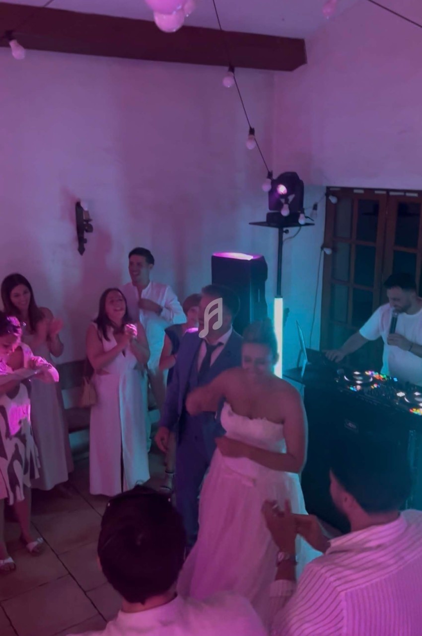 Un couple danse entouré d'invités dans une salle éclairée par des lumières colorées, avec un DJ en arrière-plan.