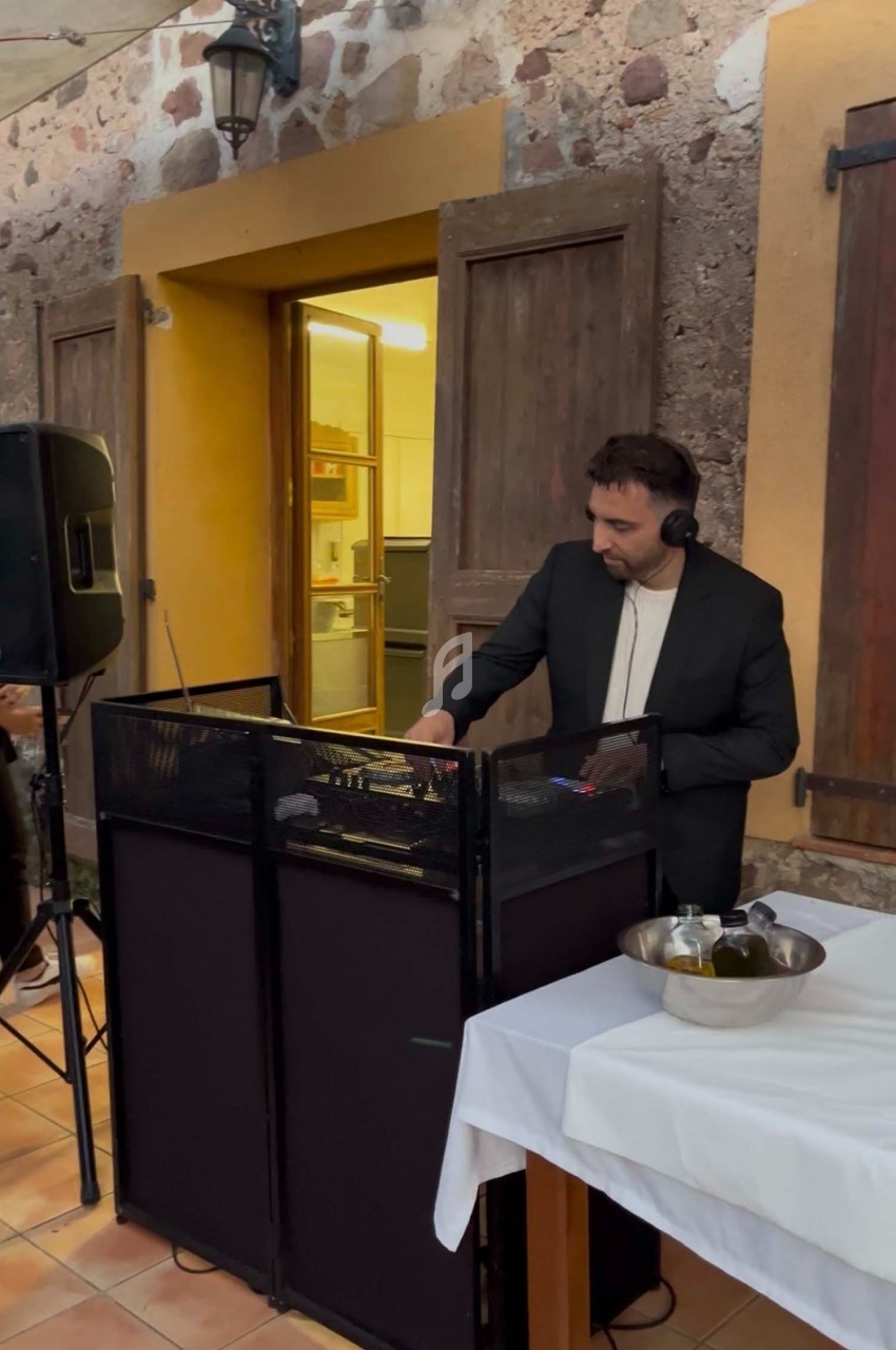 Un DJ en costume mixe de la musique sur une console dans une cour avec des murs en pierre et des tables dressées.